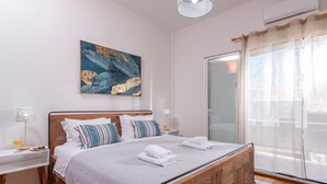 A bedroom with a wooden bed, white bedding, a bedside table with a lamp, and a large window with sheer curtains. - Sunset View Apartment (Chania)