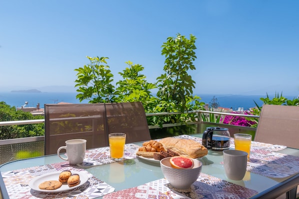 Una mesa con alimentos para el desayuno, un vaso de zumo de naranja, una cafetera y vistas al mar.