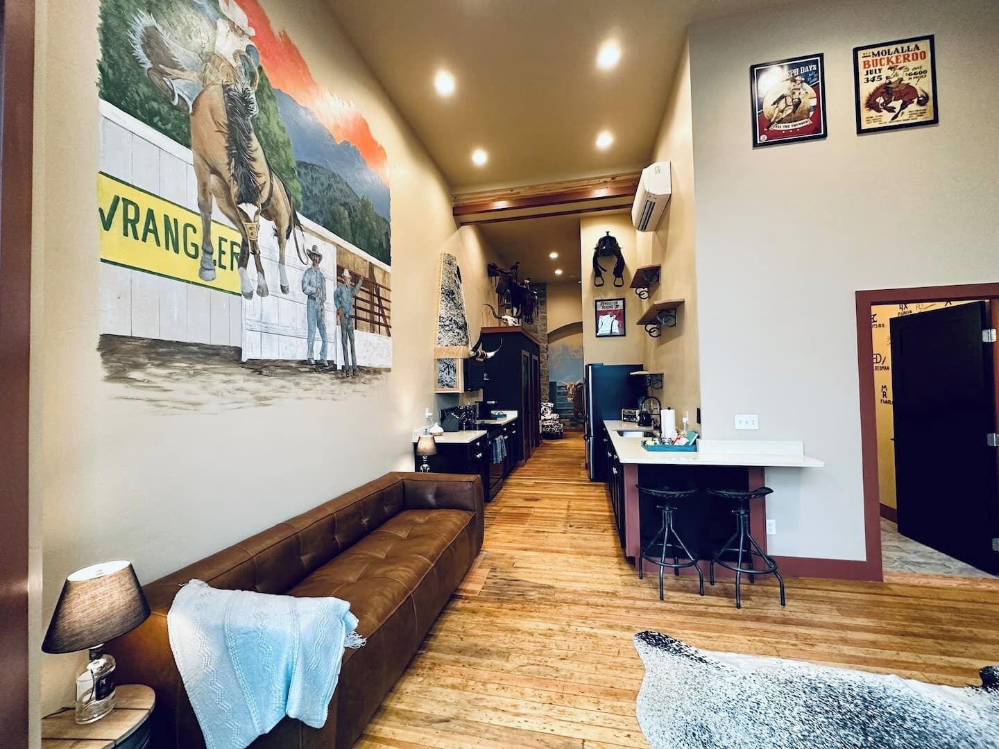 A narrow hallway with a wooden floor, a brown leather bench, a table with stools, and a wall with a mural and posters.