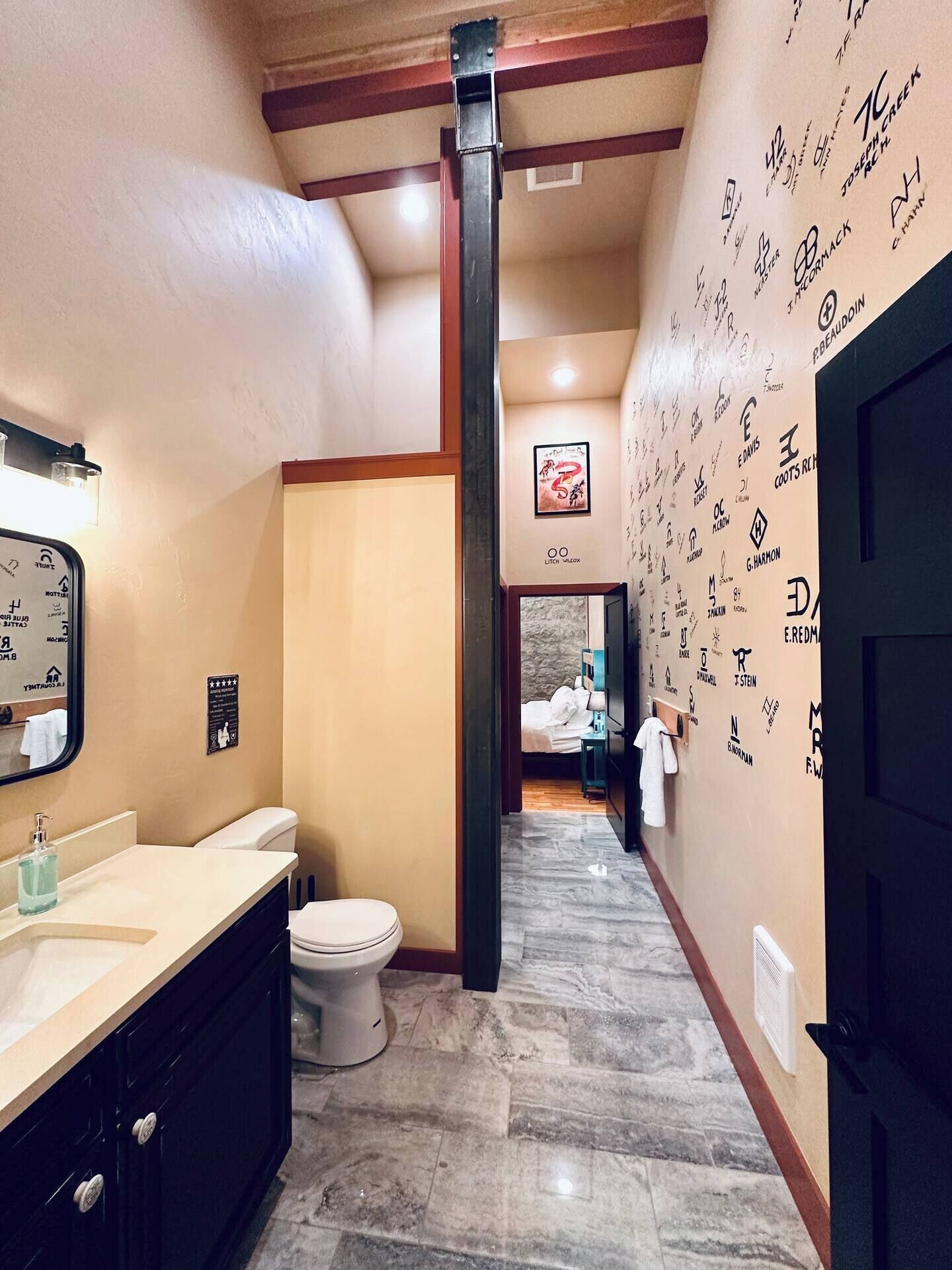 A narrow hallway with a bathroom on the left, a bedroom with a bed and a nightstand on the right, and a wall covered with various logos.