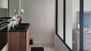 Une salle de bain moderne avec un meuble-lavabo en bois, un lavabo noir et un miroir. Un tapis noir recouvre le sol et une fenêtre offre une vue sur une chambre.