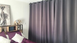 Une chambre à coucher avec un lit, une literie violette, une table de chevet avec des livres et une statuette d’éléphant décorative, et une grande fenêtre avec des rideaux gris.