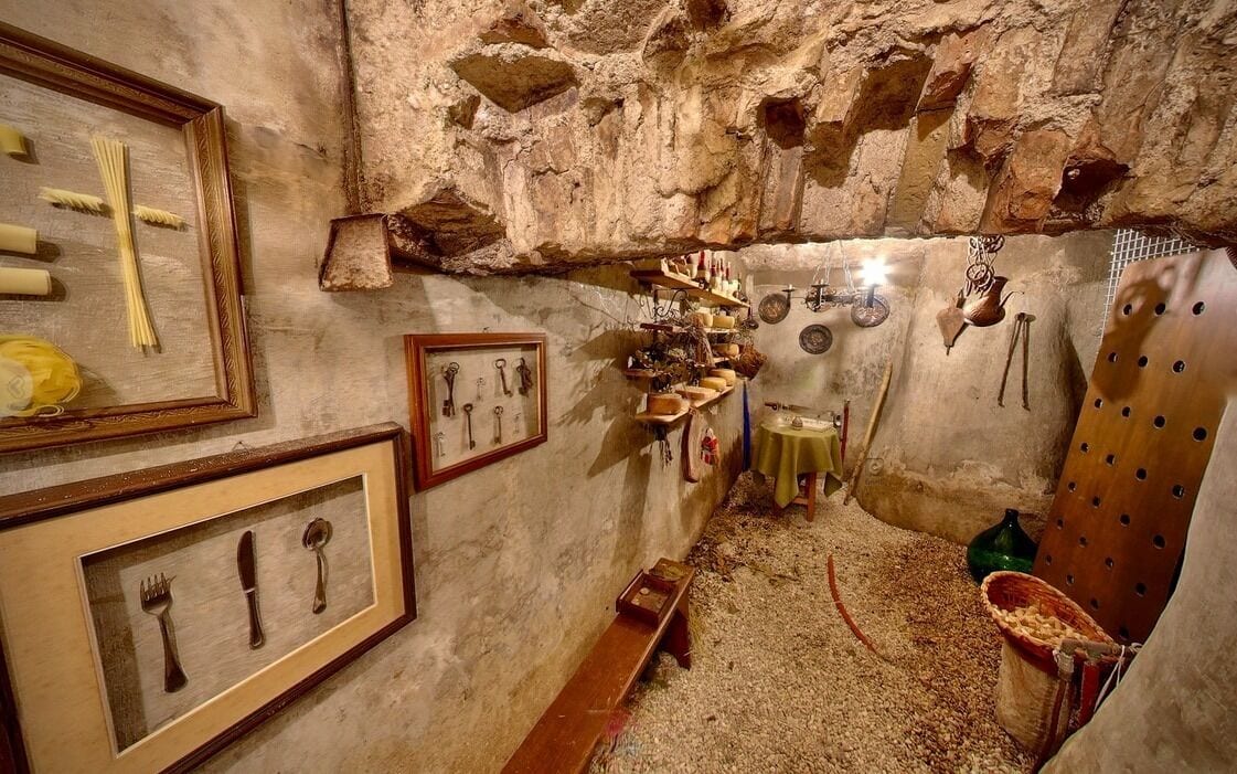 A rustic kitchen with stone walls, wooden shelves, and various kitchen utensils displayed on the walls.