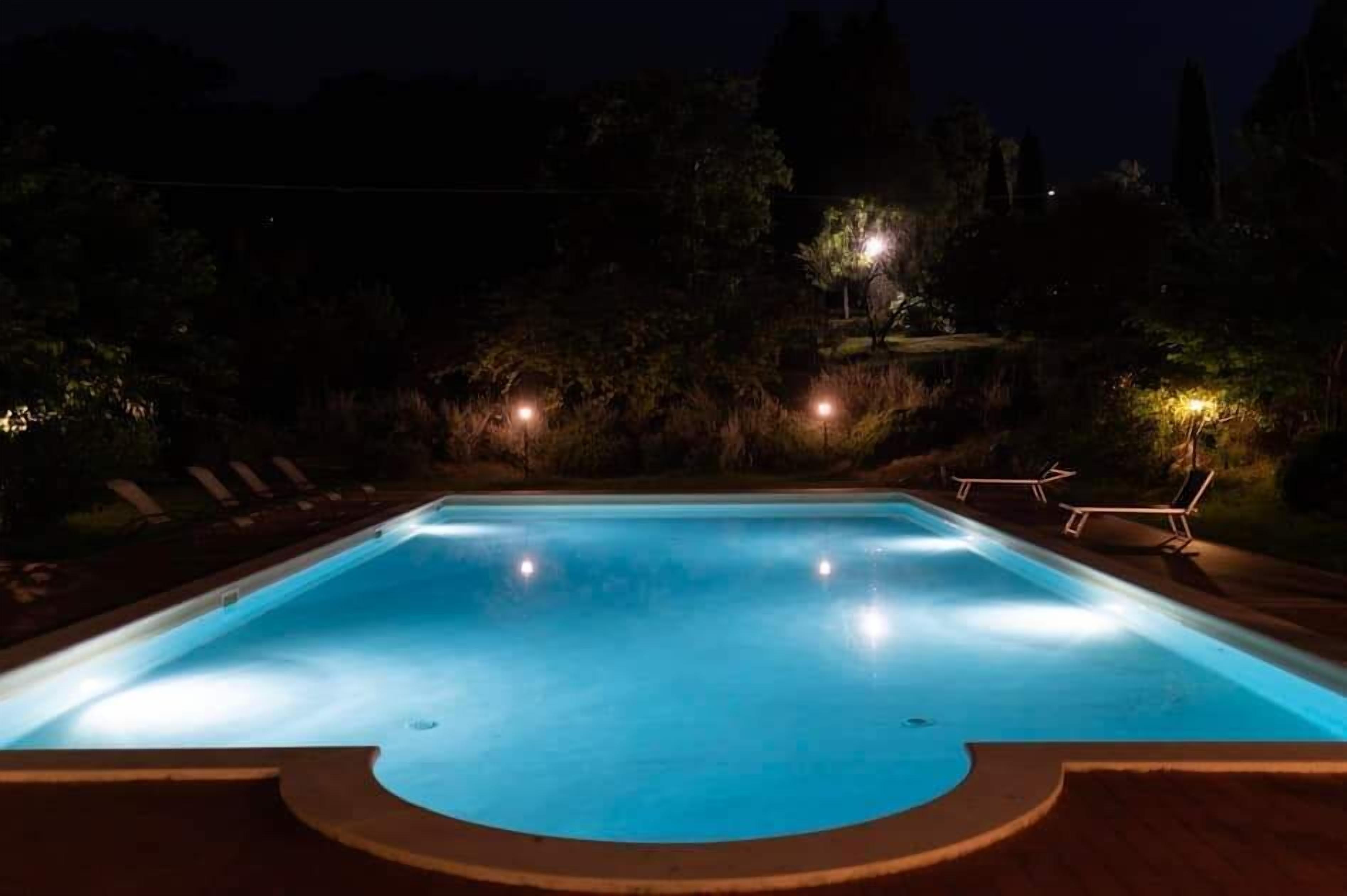 A swimming pool at night with surrounding trees and a few lounge chairs.