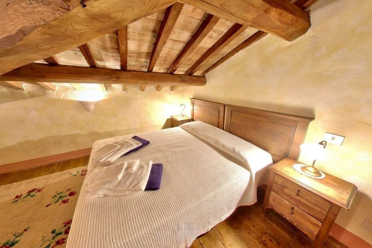Une chambre avec un plafond en bois, un lit recouvert d’une courtepointe blanche, une table de chevet avec une lampe et un tapis à motifs floraux.