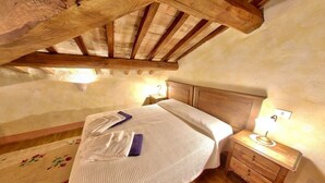 Un dormitorio con techo de madera, una cama con colcha blanca, una mesita de noche con lámpara y una alfombra estampada con motivos florales.
