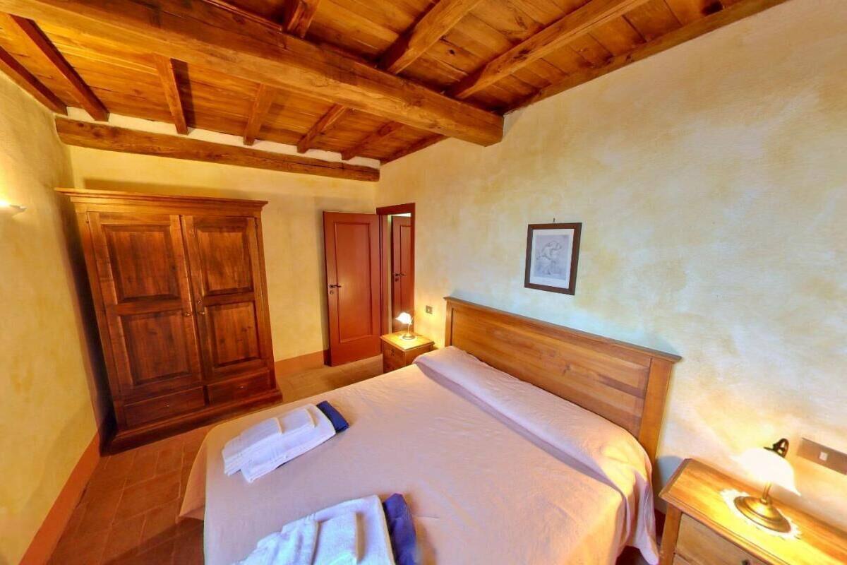 Une chambre avec un plafond en bois, un grand lit recouvert de draps blancs, une armoire en bois et une table de chevet avec une lampe.