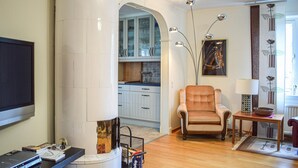 Ein Wohnzimmer mit Fernseher, einer Couch, einem Tisch mit Lampe sowie einer Küchenzeile mit Schränken und einem Fenster.