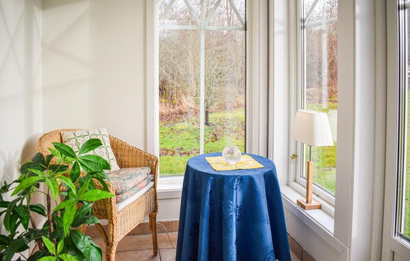 Una silla de mimbre con un cojín estampado, una mesa con mantel azul y un adorno, y una lámpara en el alféizar de una ventana.