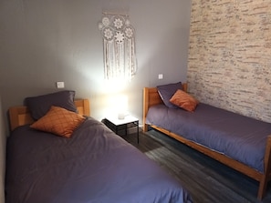 Quarto com duas camas de solteiro, cada uma com cabeceira de madeira e roupa de cama roxa. Há uma pequena mesa de cabeceira com uma luminária e um enfeite decorativo na parede acima das camas.