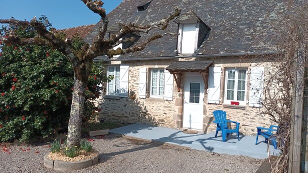 Una casa de piedra con techo inclinado, un pequeño porche con sillas azules y un árbol con flores rojas.