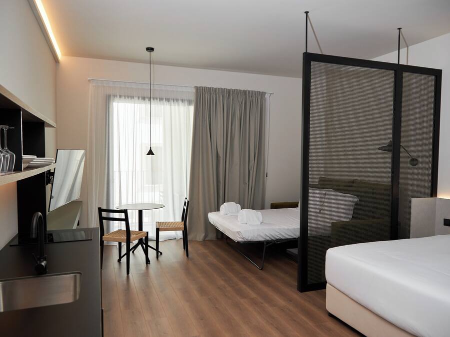 A hotel room with a bed, a desk with a sink, a chair, a table, and a large window with curtains.
