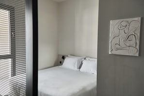 A bedroom with a bed, a wall-mounted abstract painting, and a window with a metal screen. - Alinea Primo Historic Center  (Limassol)