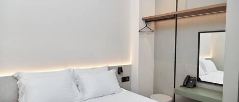 A modern hotel room with a bed, bedside table, nightstand, wall-mounted lamp, and a mirror.
