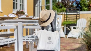 Un espacio exterior con asientos blancos, una mesa dispuesta para comer, una silla blanca y un sombrero de paja sobre una silla.