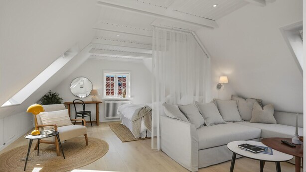 Un dormitorio tipo loft moderno con techo inclinado, un ventanal con cortinas estampadas, un sofá blanco, una mesita redonda y una silla.