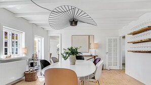 Un comedor moderno con una mesa blanca, sillas negras y doradas, una lámpara decorativa en el techo y vistas del salón comedor a través de una puerta de cristal.