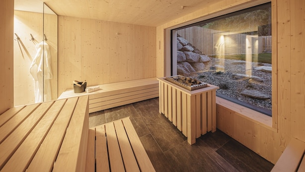 Un sauna en bois, avec un banc, une petite table ornée de pierres et une grande fenêtre donnant sur des rochers et une structure en bois.