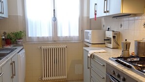 A compact kitchen with white cabinets, a stove, an oven, a microwave, and a window with curtains. - Spacious apartment with a view in the center of Montepulciano, Tuscany, Siena  (Montepulciano)