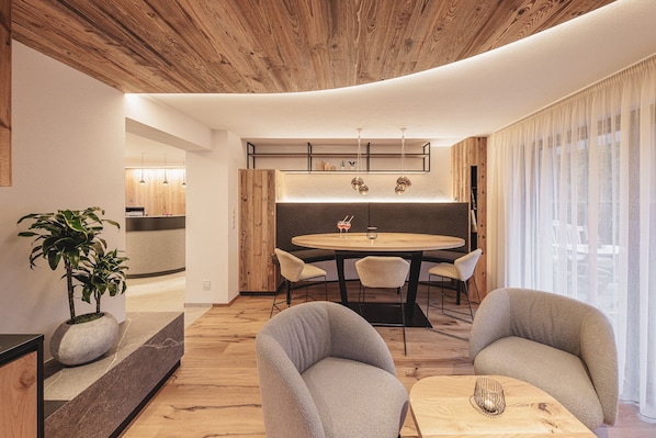A modern living room with a round dining table, two grey armchairs, a potted plant, and a wooden ceiling.