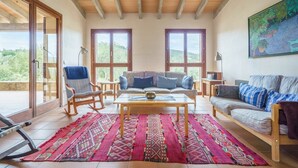 Ein geräumiges Wohnzimmer mit Holzbalkendecke, großen Fenstern, einer Sofagarnitur, einem Schaukelstuhl und einem farbenfrohen Teppich.