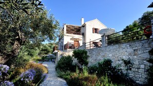 Un bâtiment aux murs de pierre, doté d’un balcon, d’un chemin bordé de pierres et entouré de verdure.
