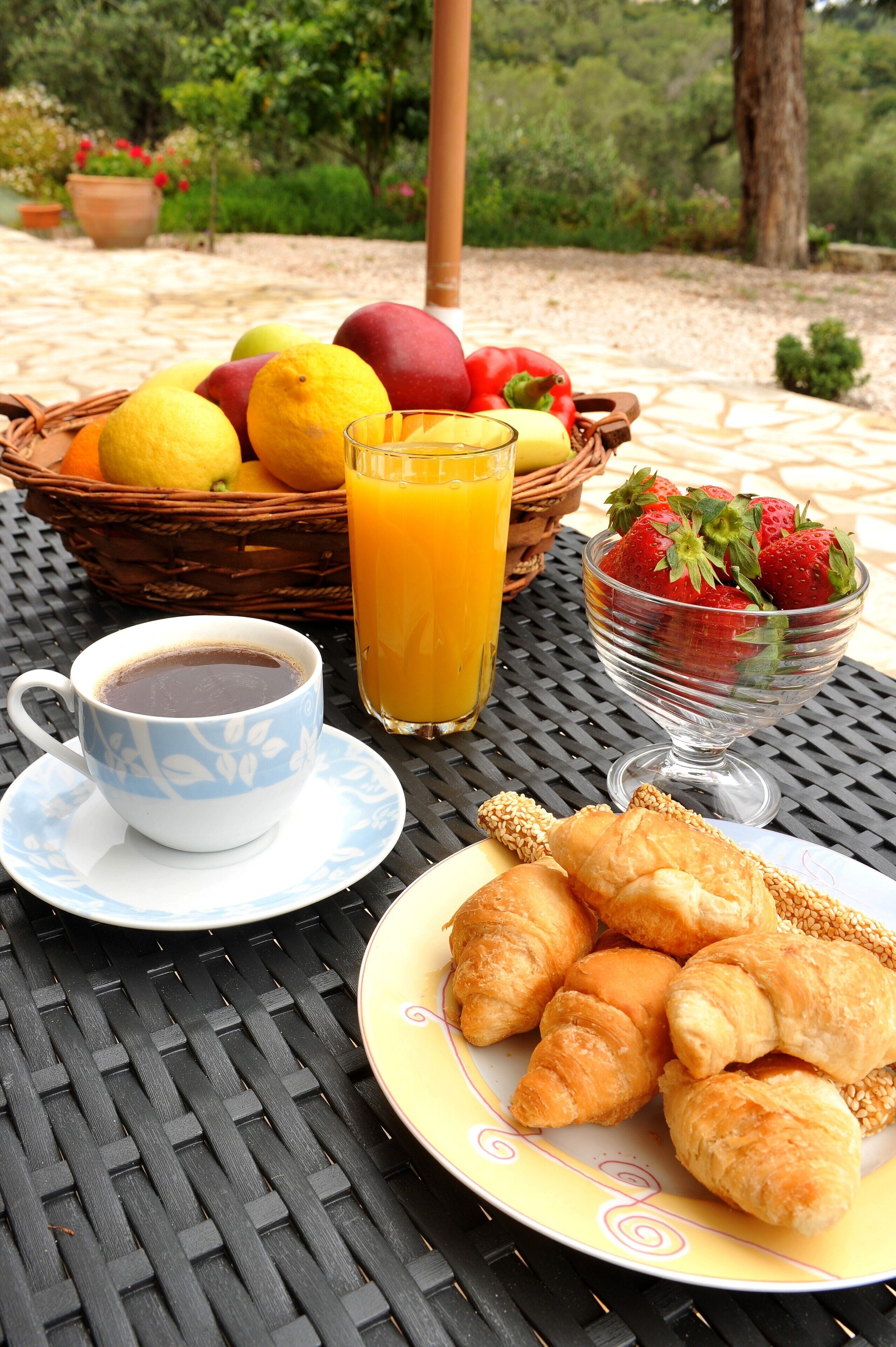 A table with a basket of fruits, a glass of orange juice, a cup of coffee, a bowl of strawberries, and a plate of croissants.