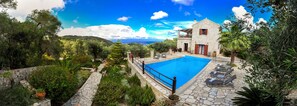 A stone house with a swimming pool, surrounded by lush greenery and a clear view of the sea.