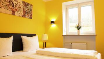A hotel room with a bed, white bedding, a black headboard, a wall-mounted lamp, and a window with a view of trees.