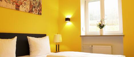 A hotel room with a bed, white bedding, a black headboard, a wall-mounted lamp, and a window with a view of trees.