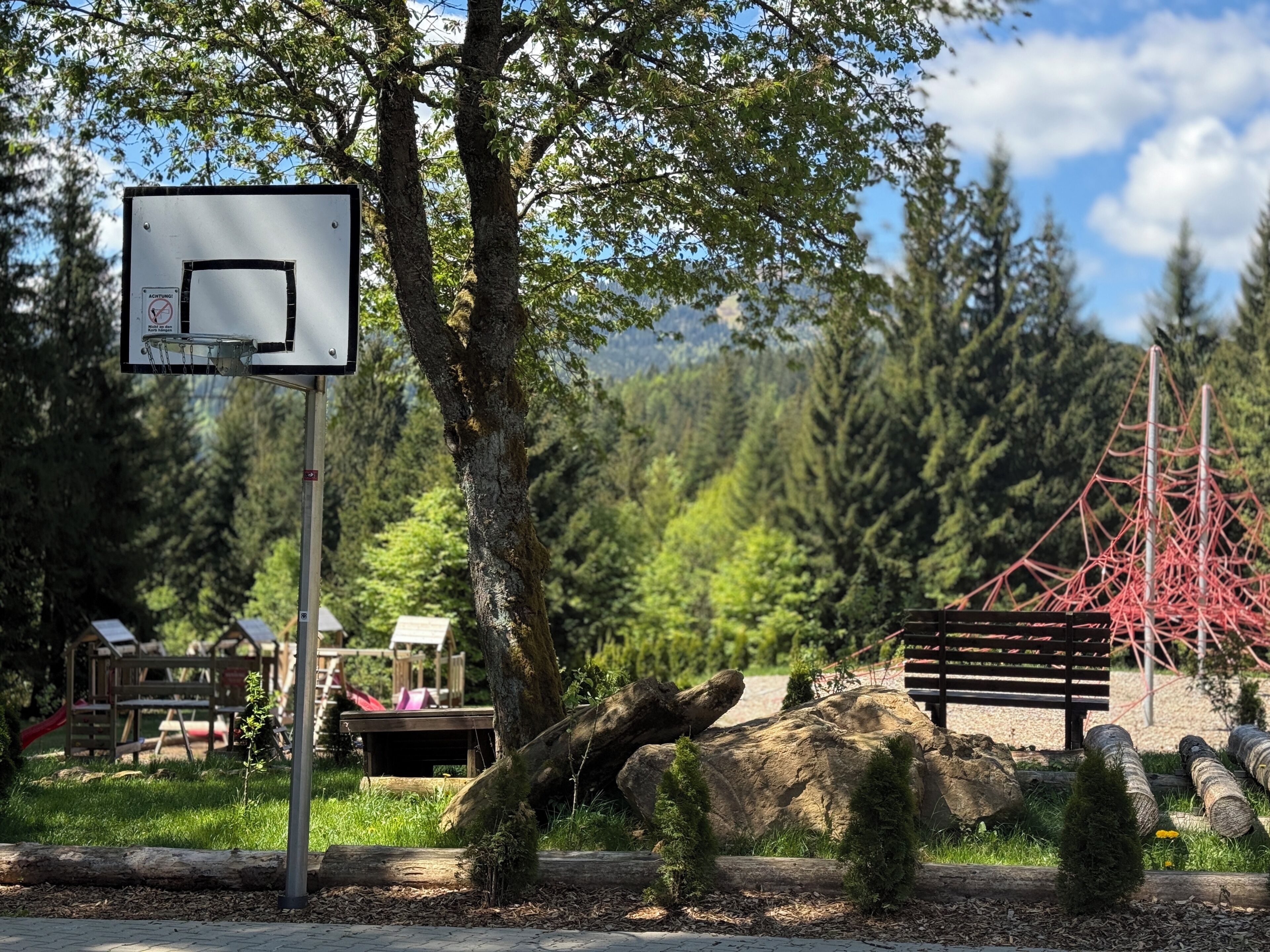 Una canasta de baloncesto, un banco de madera, un parque infantil con un tobogán y un fondo boscoso.