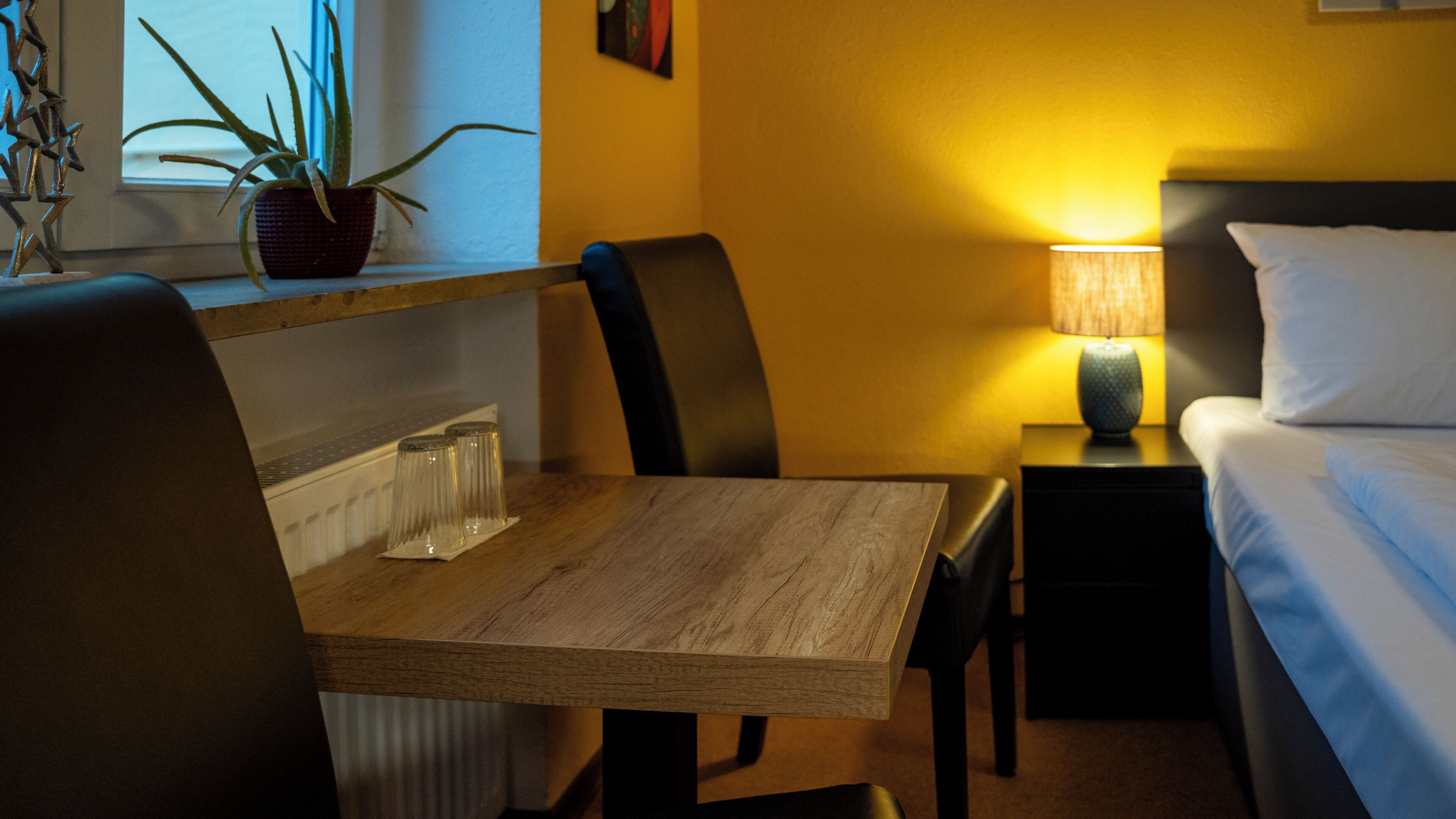 A hotel room with a wooden table, chairs, a lamp, a bed, and a potted plant.