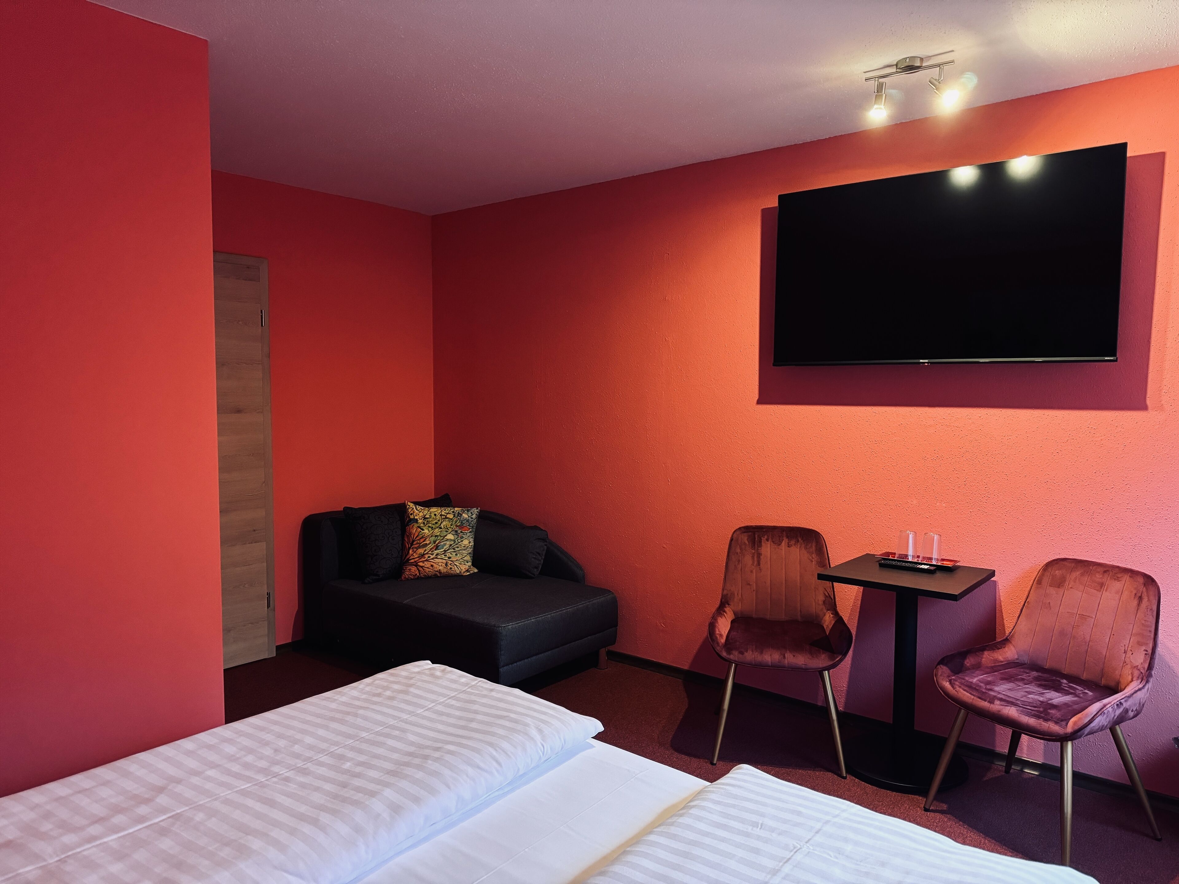 Una habitación de hotel con cama, sofá, dos sillones, una mesita y televisión montada en la pared.