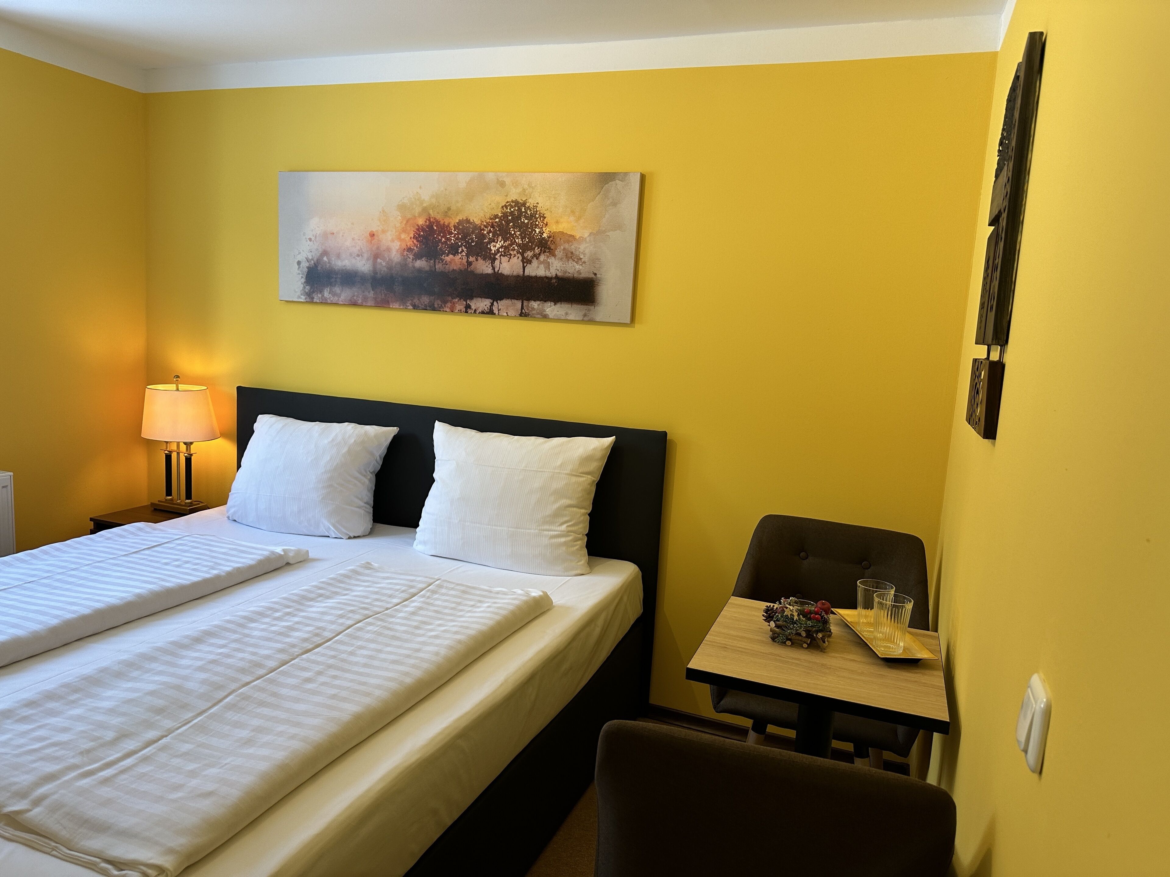 A hotel room with a bed, a nightstand with a lamp, a small table with a glass and a decorative item, and a painting on the wall.