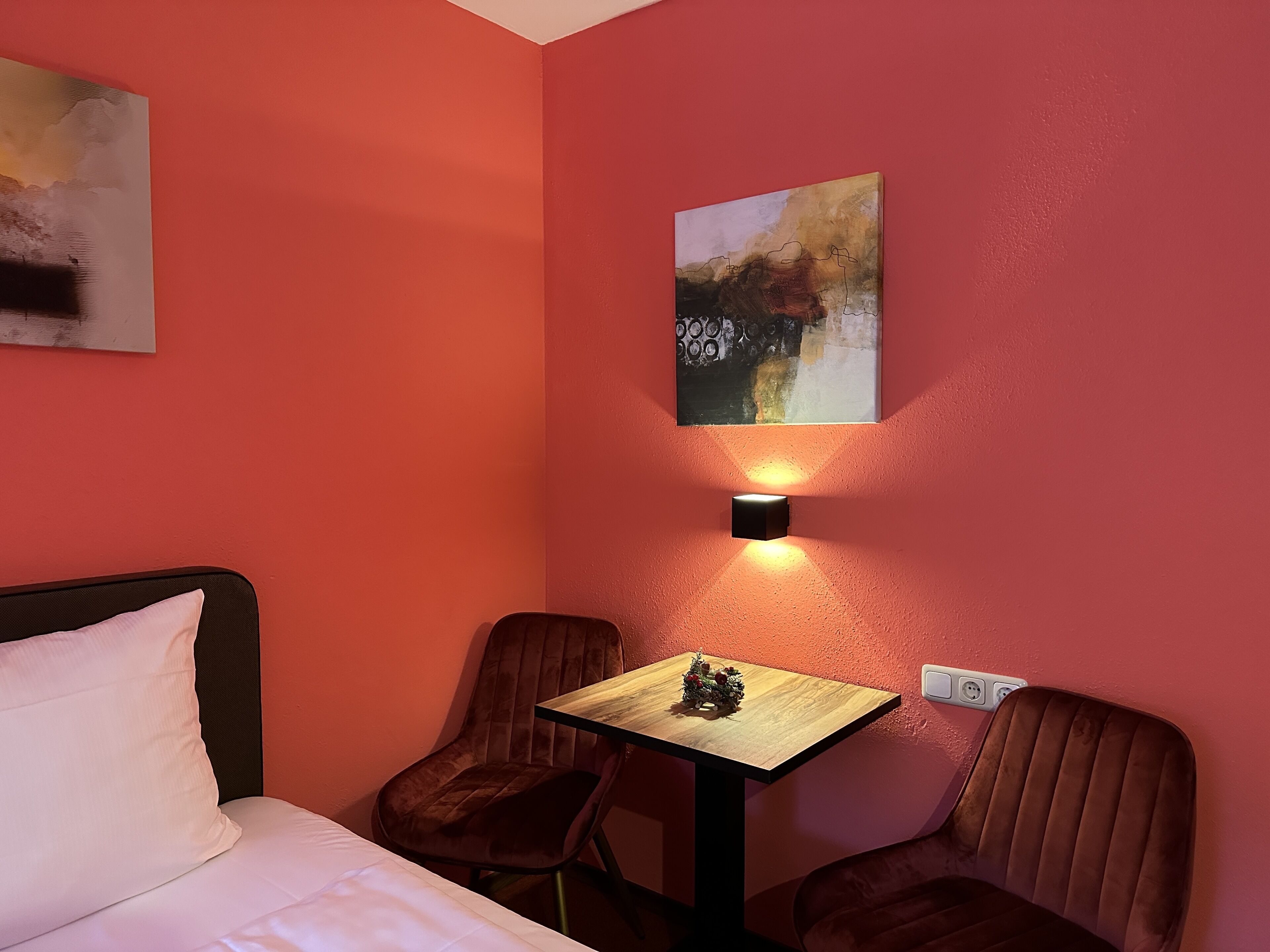 Habitació d'hotel amb un llit, dues cadires, una taula petita amb una decoració de Nadal, una làmpada penjada a la paret i un quadre.