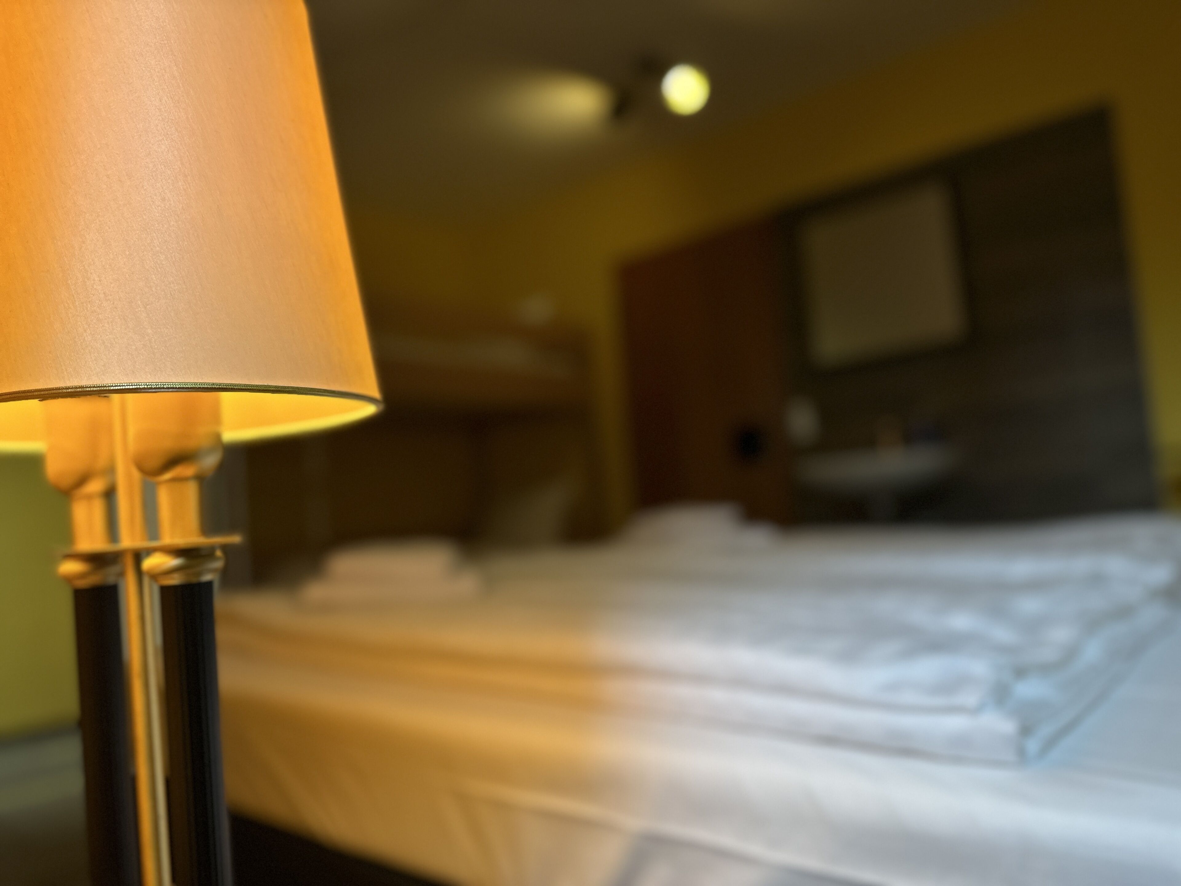 A close-up of a lamp with a beige shade, a bed with white linens, and a bathroom with a mirror and sink in the background.