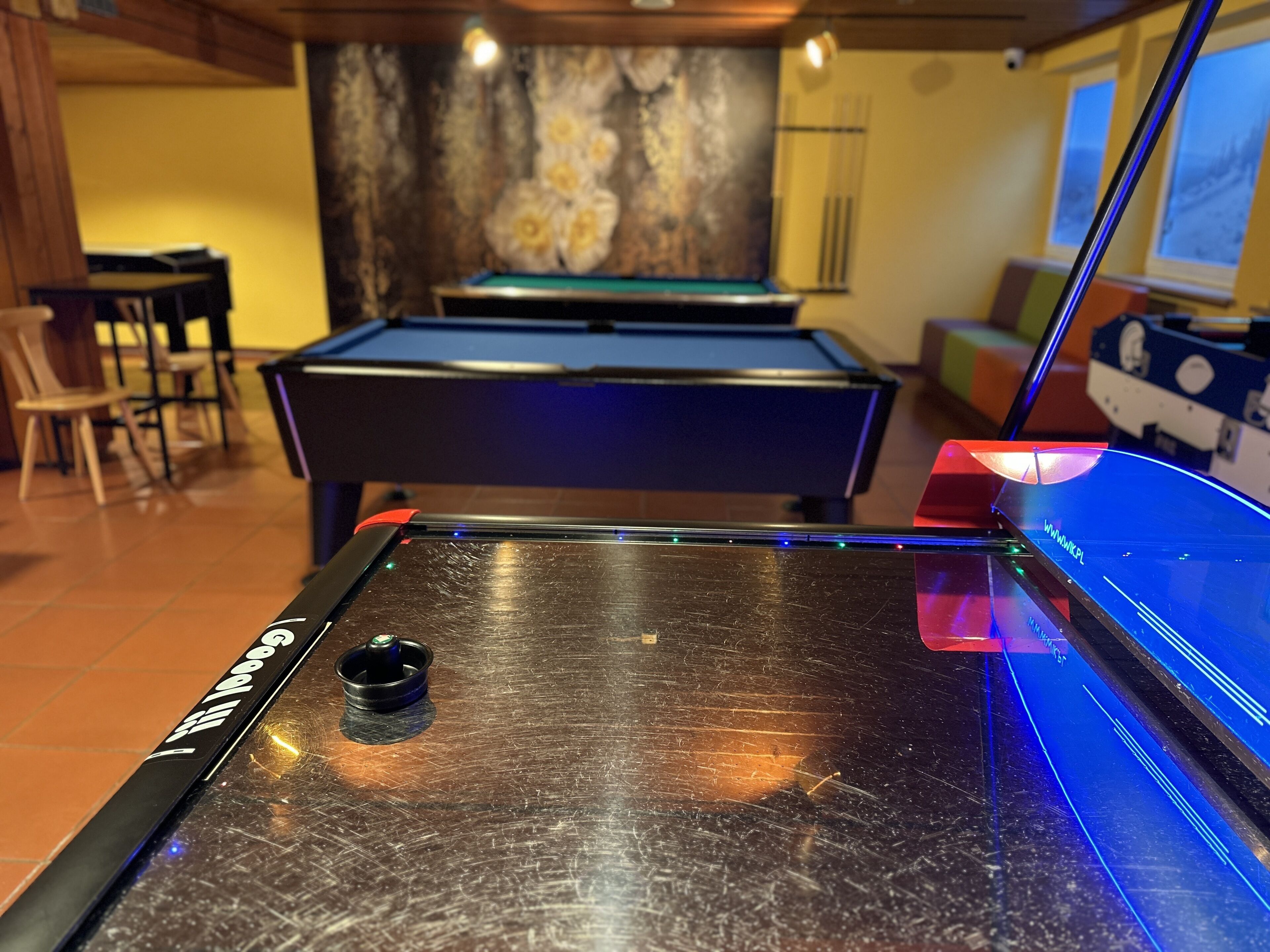 Une table de billard recouverte d’un feutre noir, présentant une éraflure visible. On y trouve un queue de billard avec un grip et un porte-billes. En arrière-plan, on aperçoit un mur orné d’un motif floral et une fenêtre.