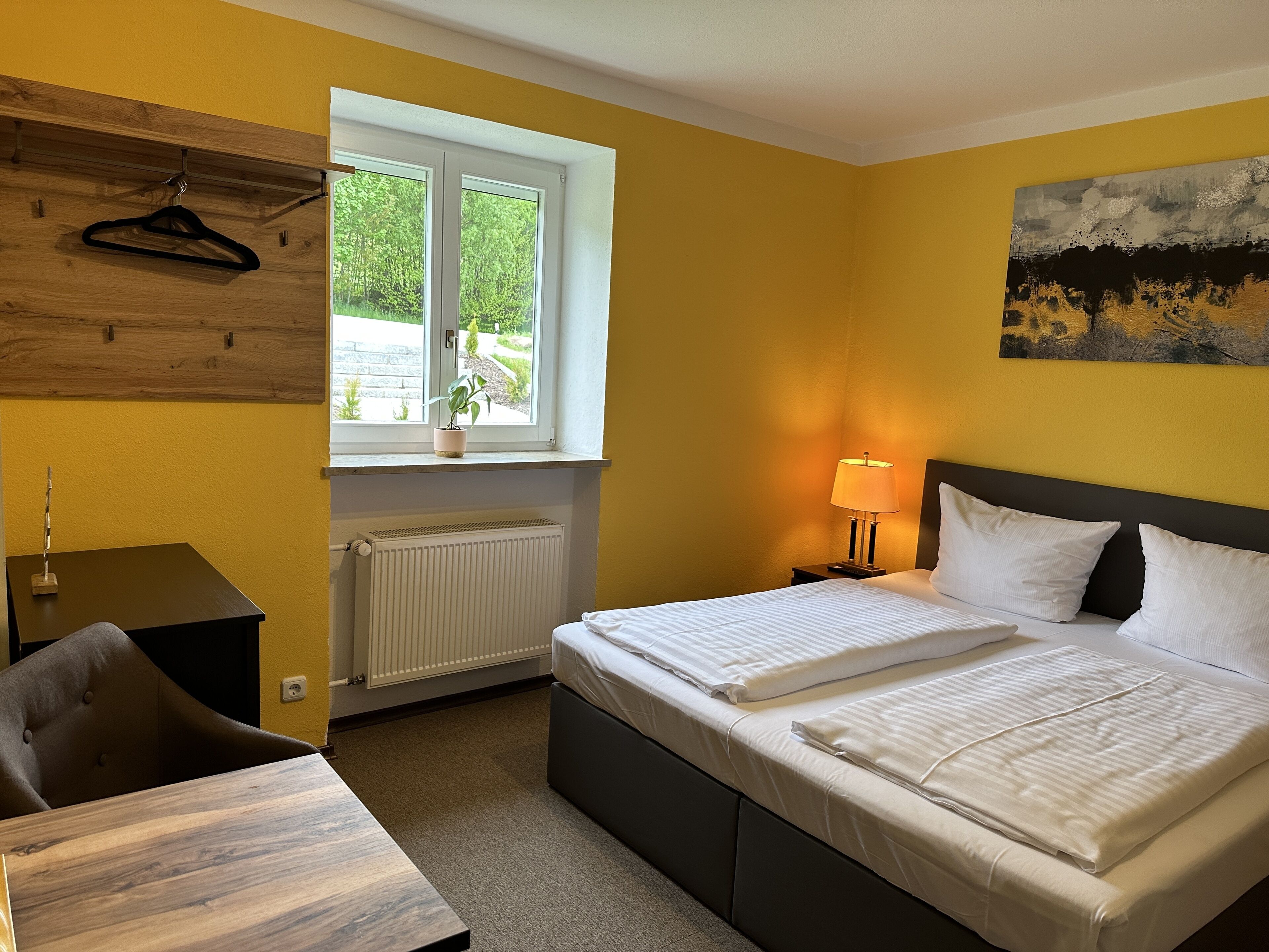 A hotel room with a bed, a desk, a chair, a radiator, a window with a view of greenery, and a painting on the wall.