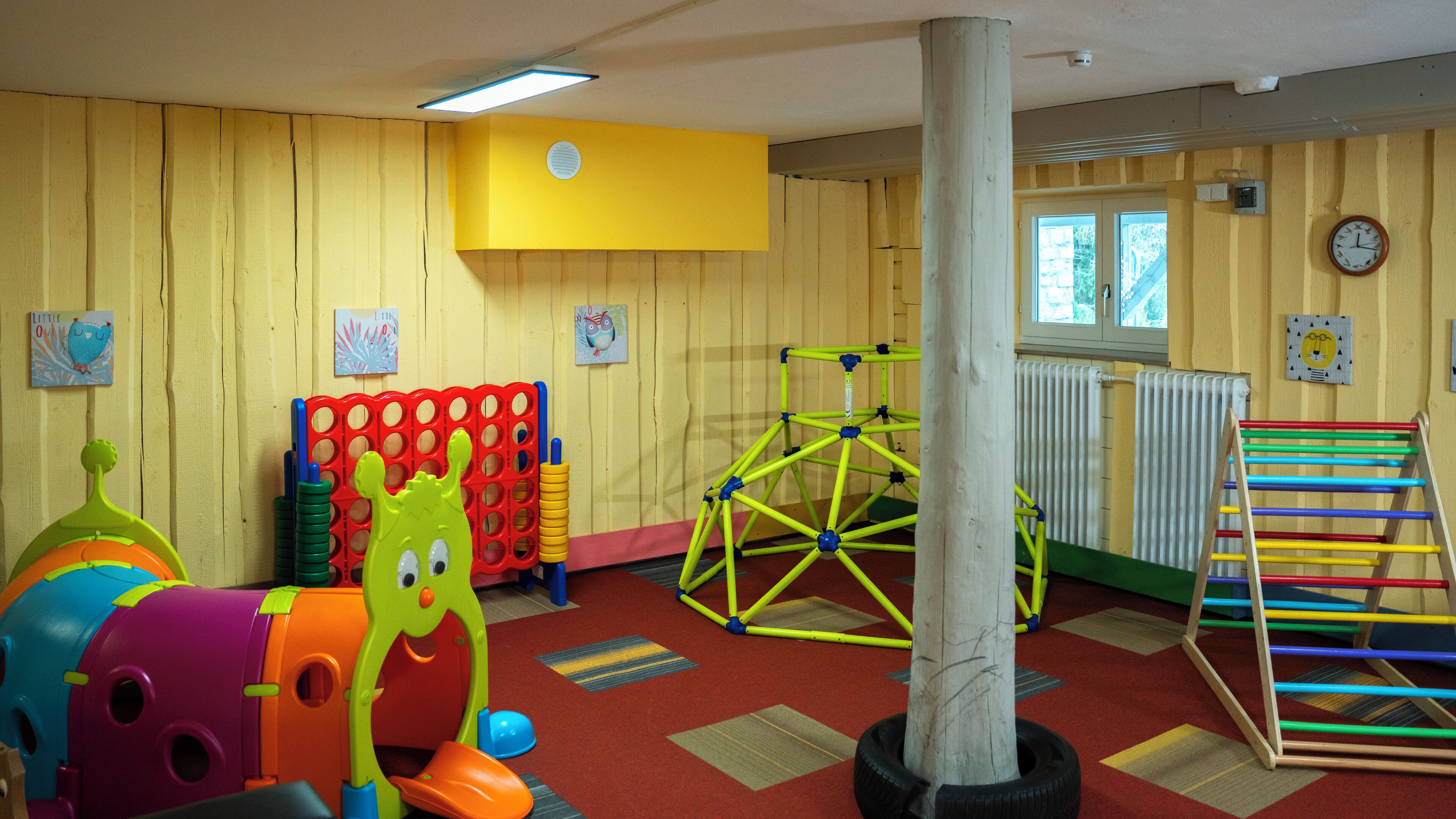Una sala de juegos con paredes de madera, una valla roja para bebés, una estructura para escalar y un tobogán colorido.
