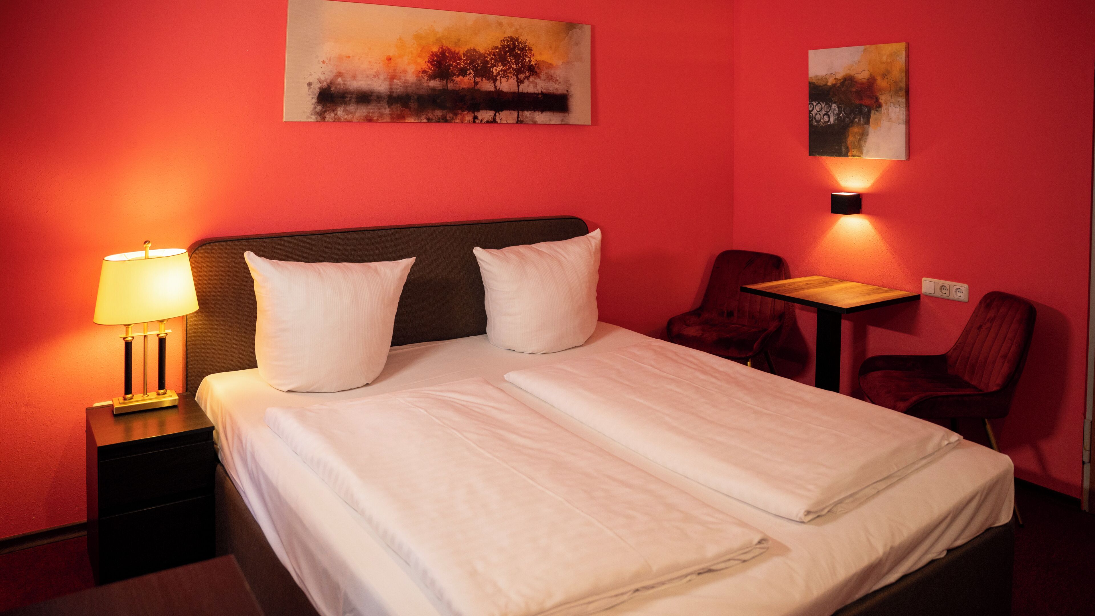 A hotel room with a bed, two pillows, a nightstand with a lamp, a chair, a small table, and two paintings on the wall.