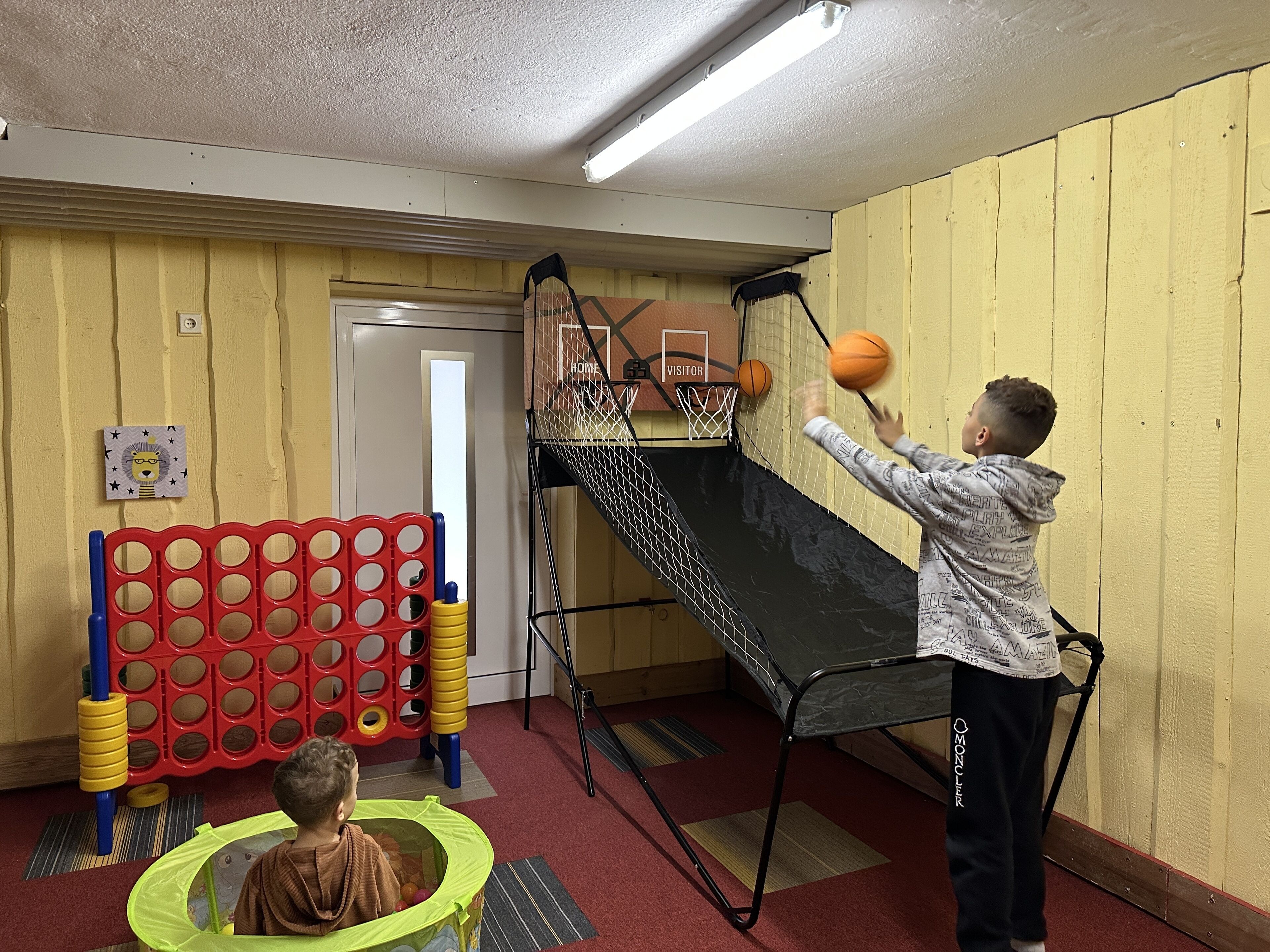 A child playing basketball in a room with a small indoor court, a red and yellow play structure, and a door labeled "Home" and "Visitor".