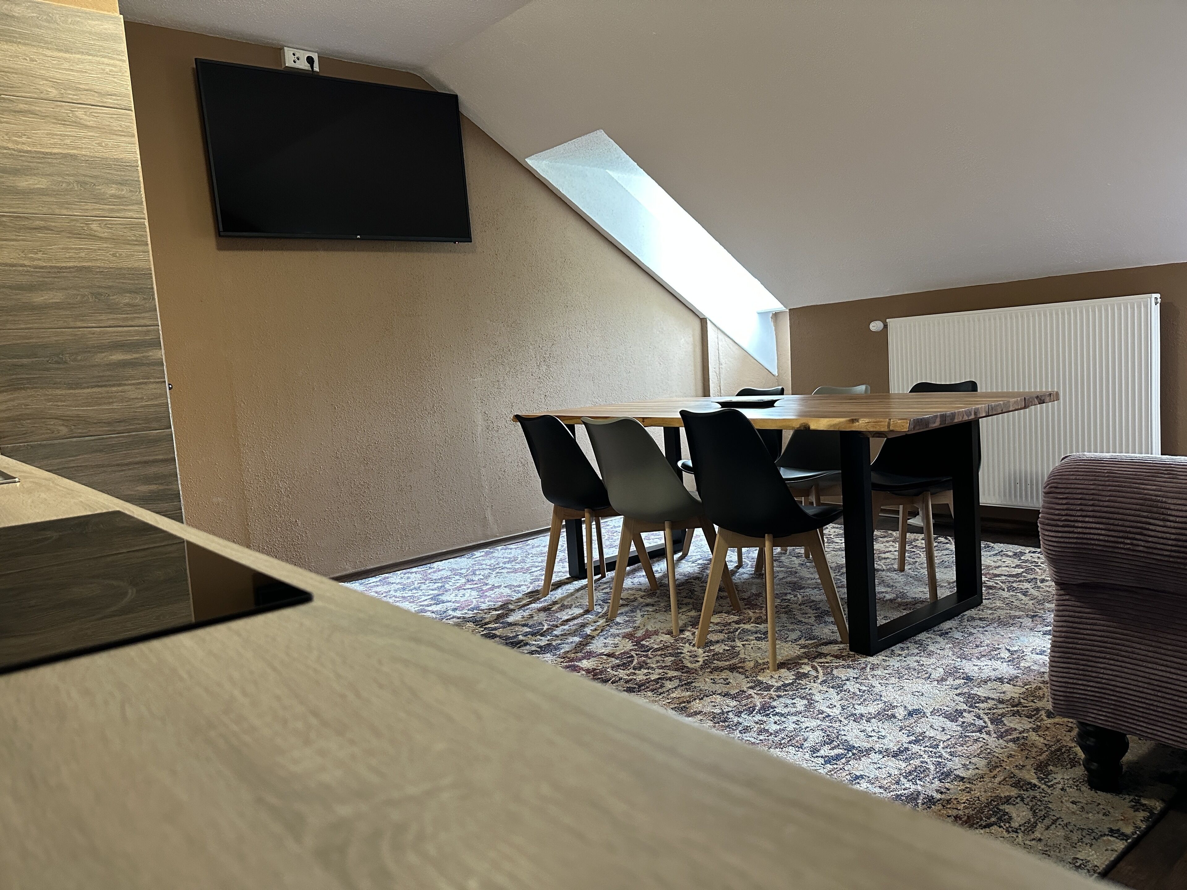 A modern dining area with a wooden table and black chairs, a flat-screen TV mounted on the wall, and a sloped ceiling with a skylight.