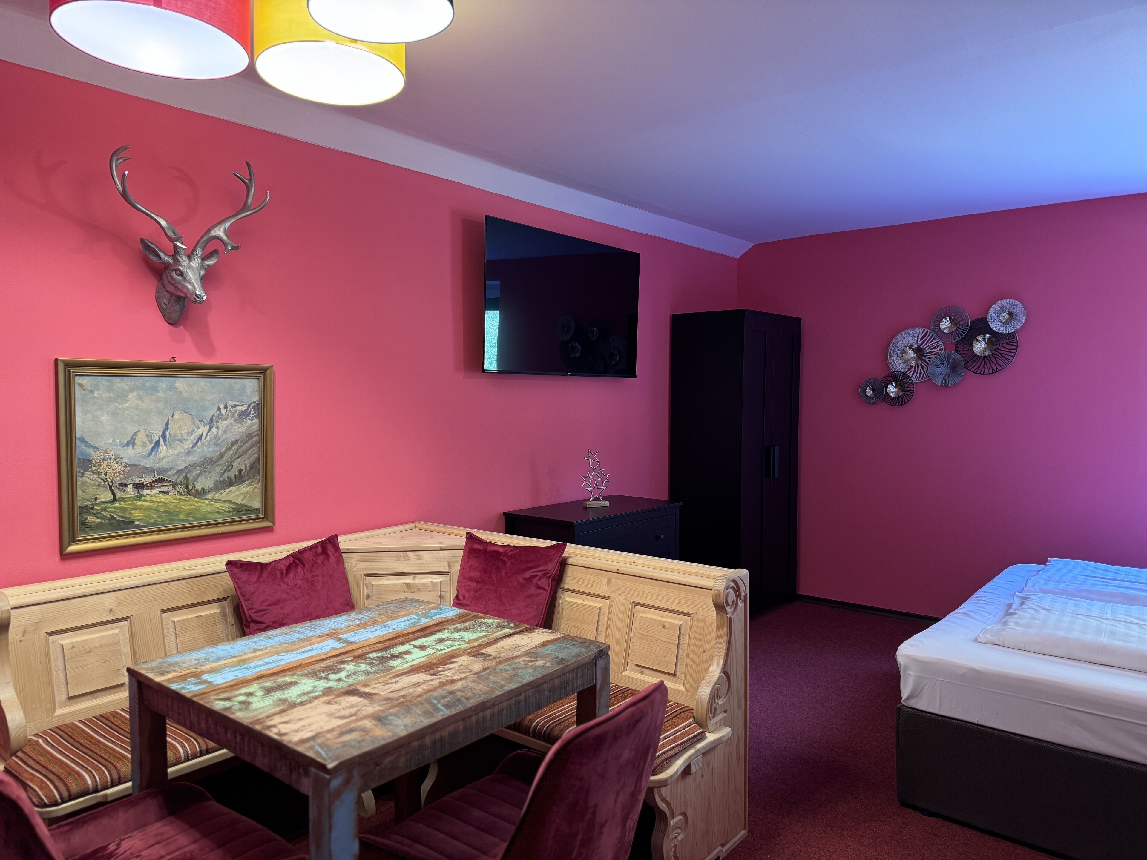 A room with a wooden table, red upholstered bench, a bed, a mounted deer head, a painting, a TV, and a cabinet.