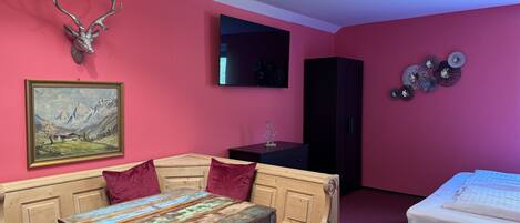 A room with a wooden table, red upholstered bench, a bed, a mounted deer head, a painting, a TV, and a cabinet.