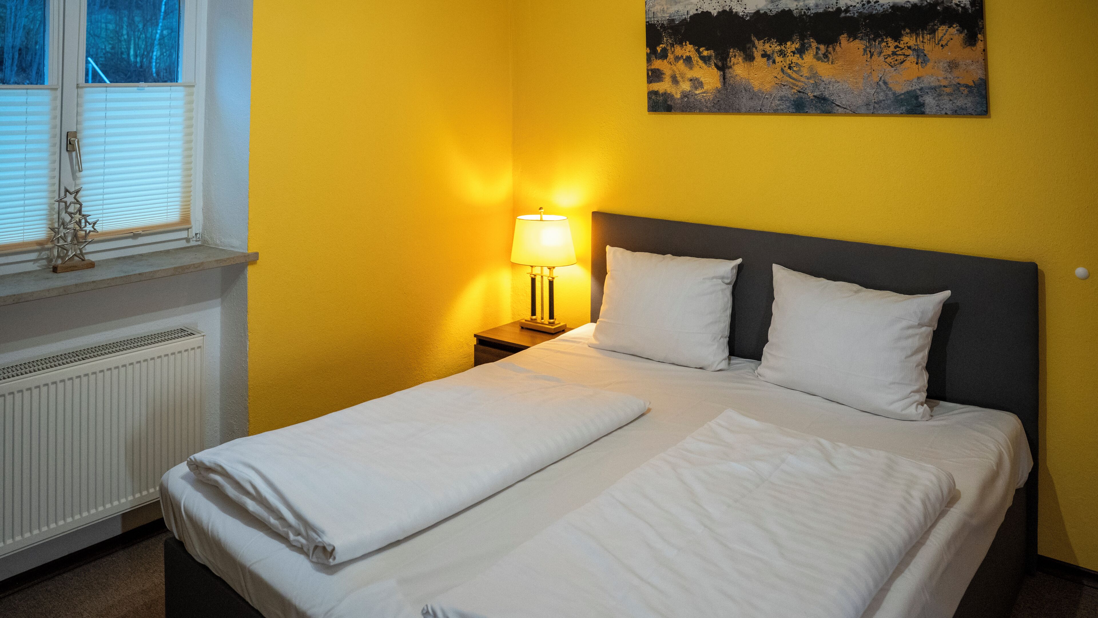 Une chambre d’hôtel avec un lit, une table de chevet, une lampe, un radiateur et une fenêtre avec des stores.