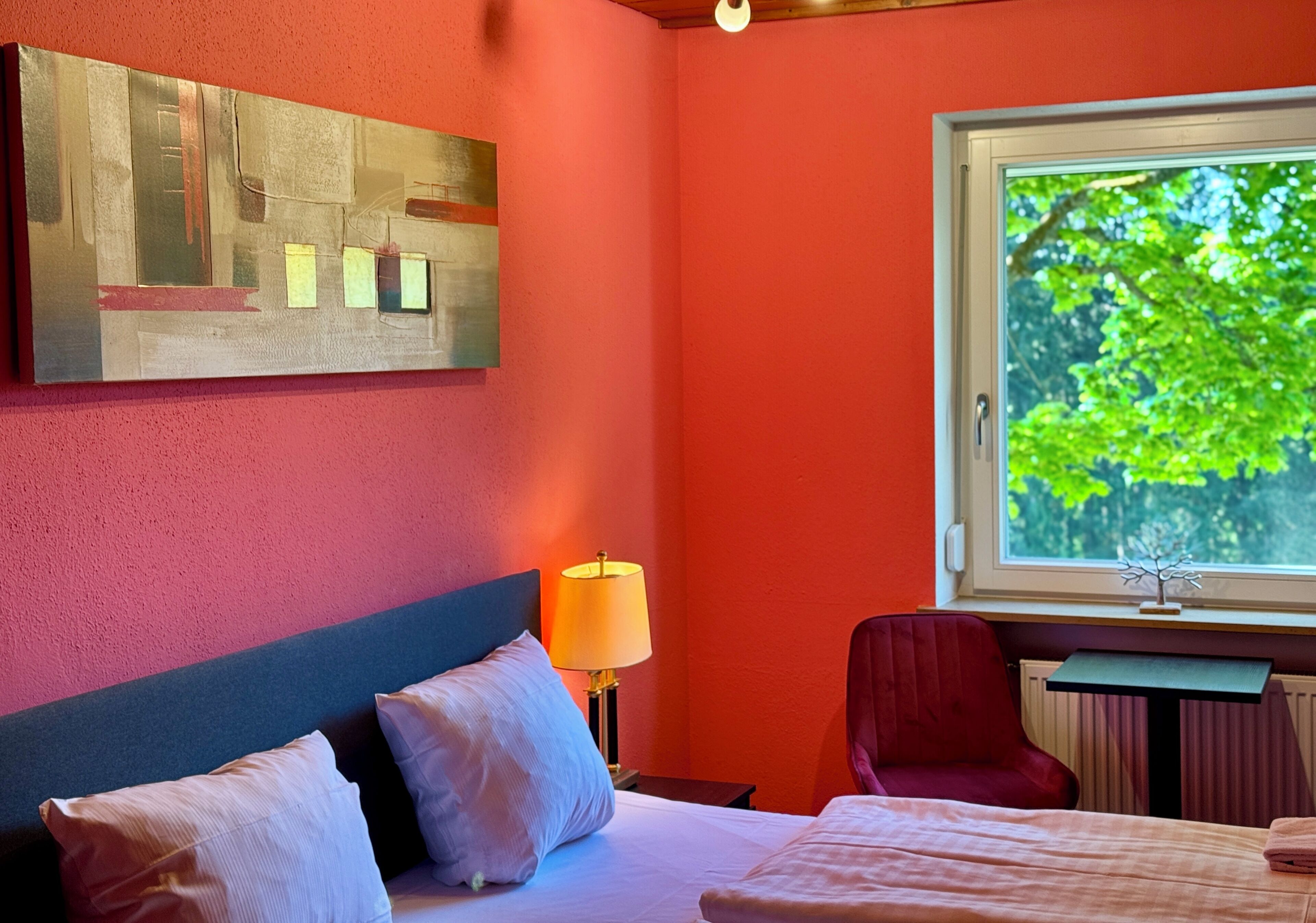 A hotel room with a bed, a desk, a chair, a window with a view of trees, and a painting on the wall.