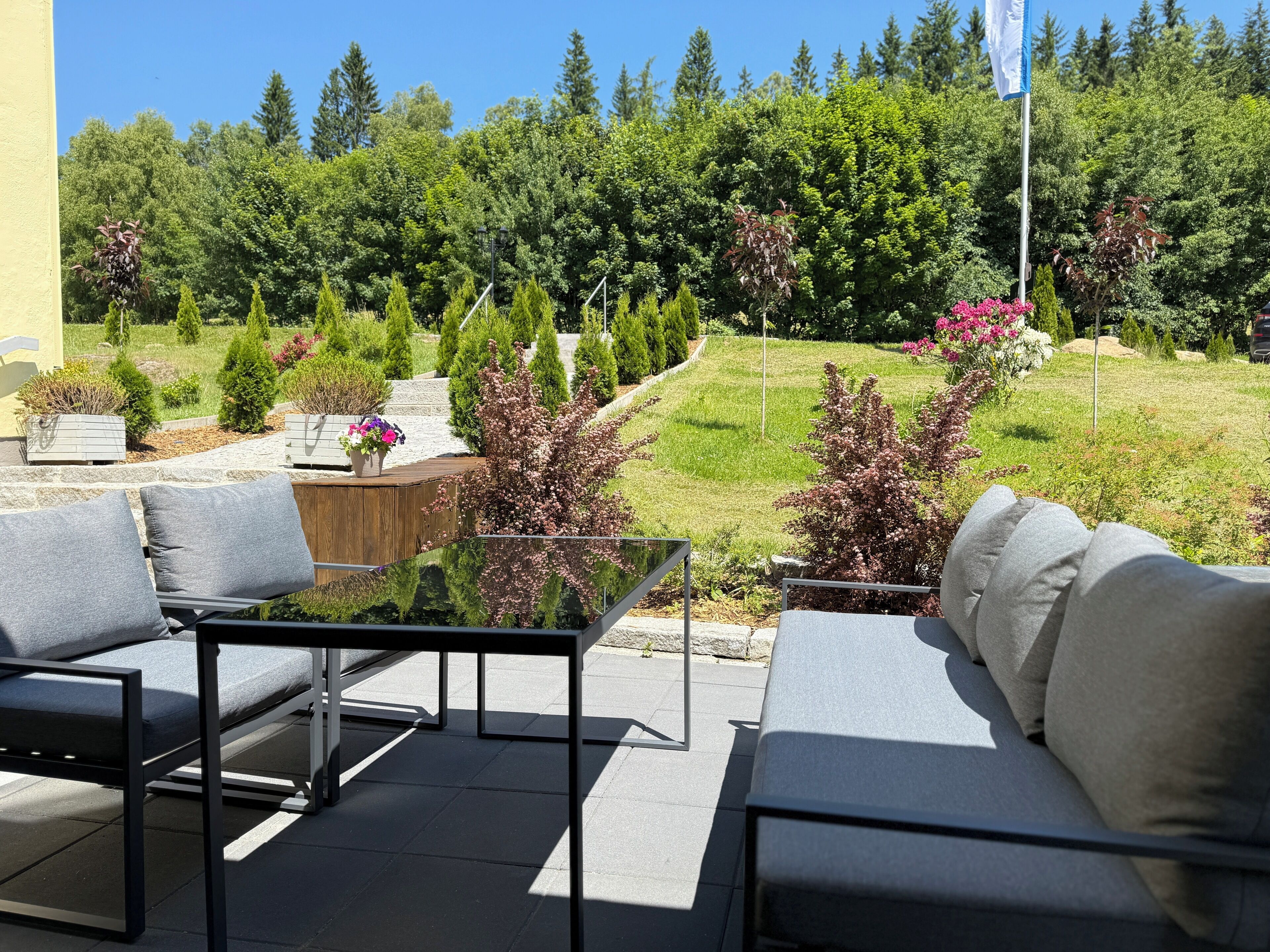 Eine Terrasse im Freien mit einem Glastisch und zwei Sofas, umgeben von einem angelegten Garten mit verschiedenen Pflanzen und einer Fahnenstange.