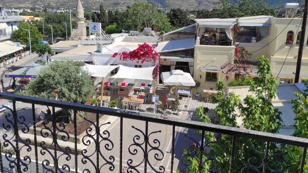 A balcony with a wrought iron railing overlooking a town with buildings, a monument, and mountains in the background.