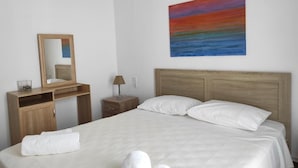 A hotel room with a bed, a wooden desk, a nightstand, a lamp, a mirror, and a painting on the wall.
