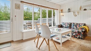 A room with a wooden floor, white walls, a floral sofa, a white table, and a window overlooking a deck.